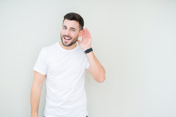 Young handsome man wearing casual white t-shirt over isolated background smiling with hand over ear listening an hearing to rumor or gossip. Deafness concept.