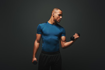 Achieving best results. Dark skinned sportsman working out with dumbbells over dark background