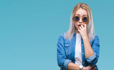 Young blonde woman wearing sunglasses over isolated background looking stressed and nervous with hands on mouth biting nails. Anxiety problem.