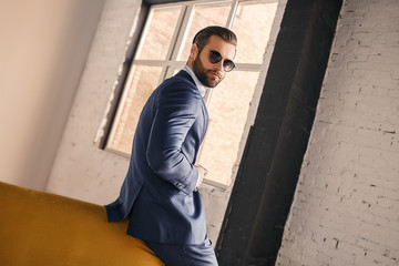 Being successful. Portrait of pleasant and young businessman in sunglasses and stylish suit who is standing at office