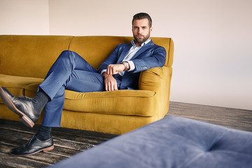 Good looking young businessman in a stylish suit is relaxing on sofa and looking at camera