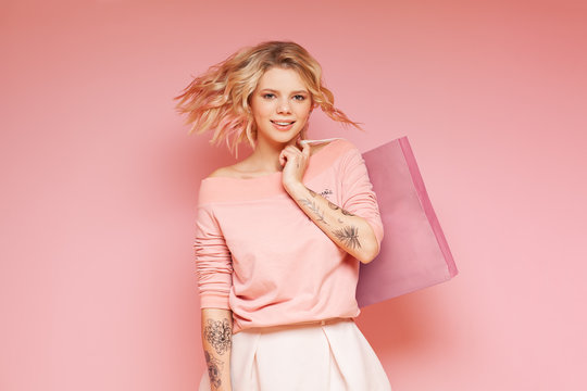 Young Student Hipster Woman With Colored Flying Hairs And Tattoo Holding Shop Bag. Isolated On The Pink Background