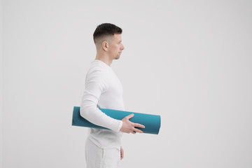 A male teacher and trainer in white clothes holding a yellow-blue yoga mat in his hands, preparing for a fitness class.