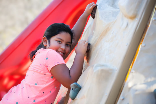 Young latino girl climbing a playground wall and smiling.