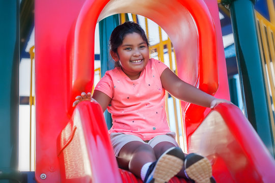 A pretty young hispanic girl with a cute smile looks relaxed as she prepares to slide down a playground slide.