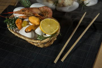 Hot king prawns, mussels and fish with lemon lie on white heated stones with steam. Serving food on a black wooden sushi rug with sushi chopsticks
