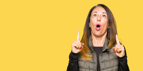 Beautiful middle age woman wearing winter vest amazed and surprised looking up and pointing with fingers and raised arms.