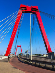 Cyclist crossing the Willemsbrug bridge spanning the Nieuwe Maas river in Rotterdam, the Netherlands. Red bridge pylons and cables against blue sky