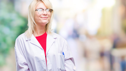 Young beautiful blonde professional woman wearing white coat over isolated background looking away to side with smile on face, natural expression. Laughing confident.