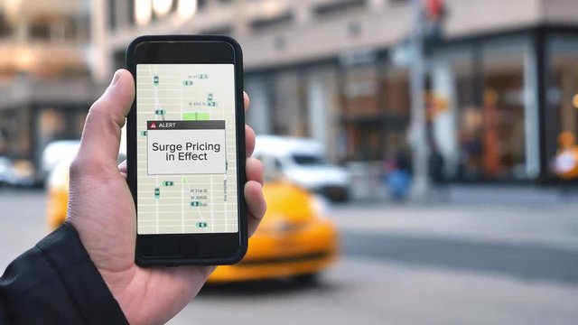 A man uses a ride-sharing app on his cellular phone to call a driver and notices a surge pricing pop-up warning.	 	