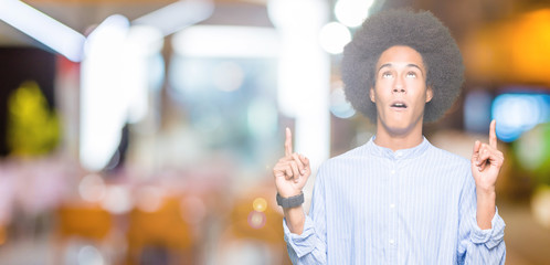 Young african american man with afro hair amazed and surprised looking up and pointing with fingers and raised arms.