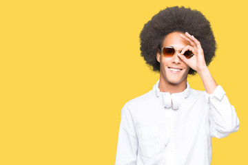 Young african american man with afro hair wearing sunglasses and headphones doing ok gesture with hand smiling, eye looking through fingers with happy face.