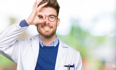 Young handsome scientist man wearing glasses doing ok gesture with hand smiling, eye looking through fingers with happy face.