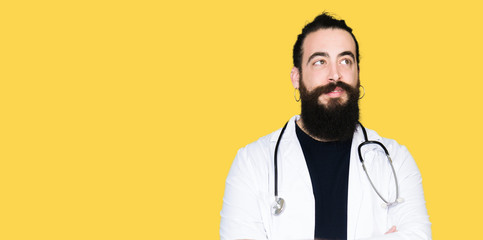 Doctor with long hair wearing medical coat and stethoscope smiling looking side and staring away thinking.