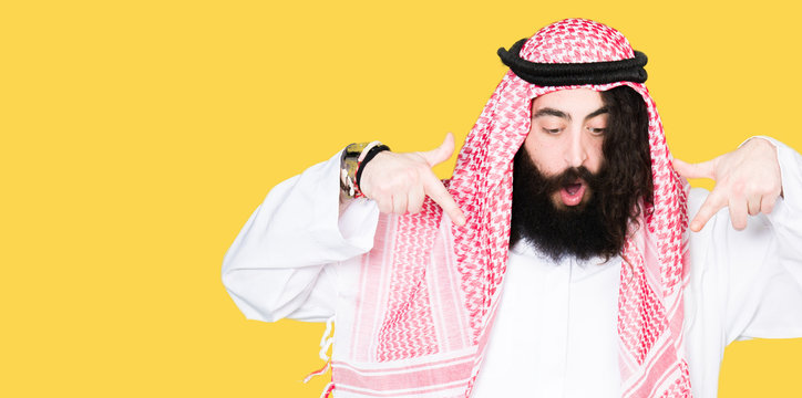 Arabian business man with long hair wearing traditional keffiyeh scarf Pointing down with fingers showing advertisement, surprised face and open mouth
