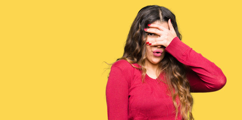 Young beautiful woman wearing red sweater peeking in shock covering face and eyes with hand, looking through fingers with embarrassed expression.