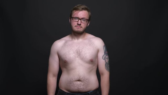 Portrait of young naked overweight man watching sadly into camera on black background.