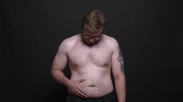 Portrait of young naked overweight man watching sadly into camera and touching his stomach on black background.