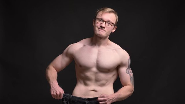 Portrait of naked young muscular man in glasses happily showing the results of losing weight into camera on black background.
