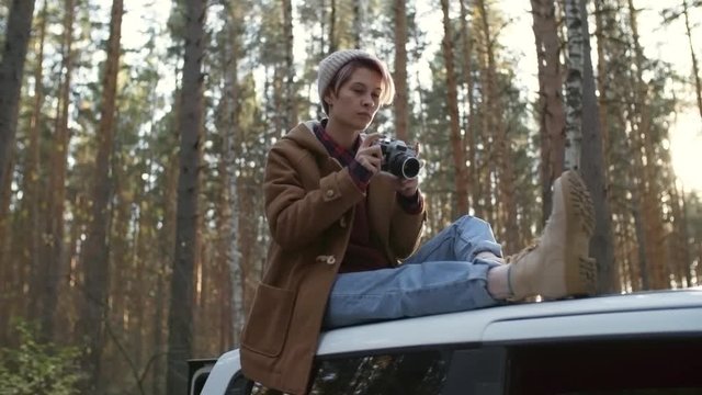 Young Caucasian woman in beanie sitting on car roof on forest road and using camera to take photos of nature in autumn