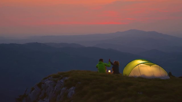SLOW MOTION: Cheerful couple sitting in the grass high fives while watching the sunset during a fun camping adventure in the serene mountains. Active man and woman on a romantic outdoor date in Alps.