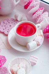 A white tea cup with pink scented tea and saucer sits on a white background with colourful paper bunting and marshmallow sweets