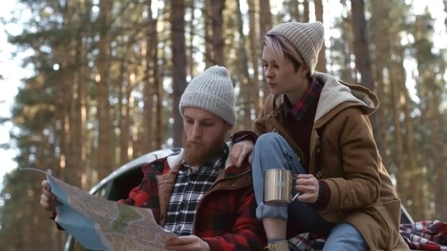 Medium shot of young Caucasian couple in beanies relaxing during car trip and choosing places to visit using paper map
