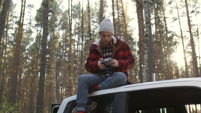 Bearded young man in beanie sitting on car roof and using camera to take photos of forest nature