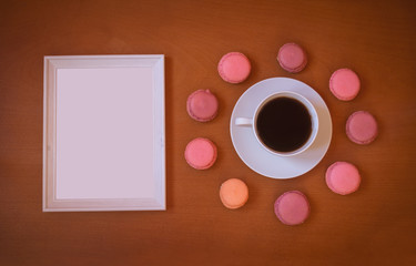 White frame and cup of coffee with colorful macaroons on wooden background.