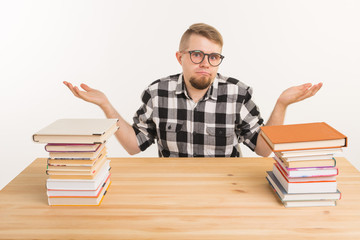 Exam, knowledge and education concept - confused student man sitting at the table with mountains of books