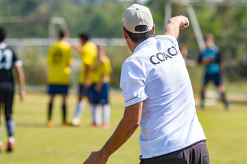 Back of football coach wearing white COACH shirt at an outdoor sport field coaching his team during a game