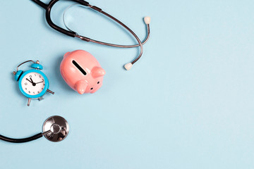 Piggy bank with stethoscope and alarm clock on blue background. Top view with copy space.