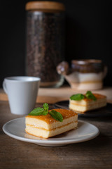 layer vanilla flavoured cake place on white plate on the wooden table there are white cup and black plate and coffee bean in glass bottles and watering ceramic place backside.