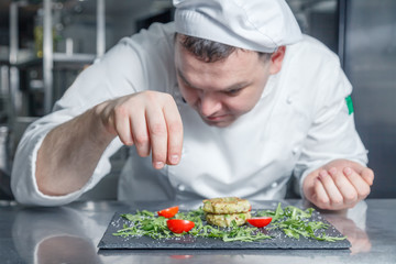 Closeup portrait caucasian man chef in restaurant kitchen prepared, decorate and salt healthy diet dish, zucchini pancakes with arugula, cherry tomato on a black slate rectangular plate