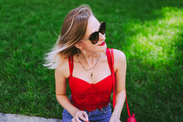 Beautiful happy girl in red tops, blue jeans and sunglasses sits in a park on the grass. Outdoor portrait of young attractive woman.