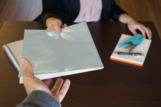 Woman is handing over a white file in direction camera while sitting at the desk in the office.