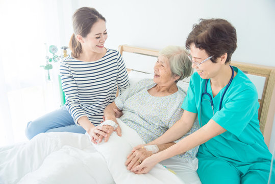 Doctor talking to happy elderly female patient lying in bed while patient's daughter sitting beside her on bed in hospital