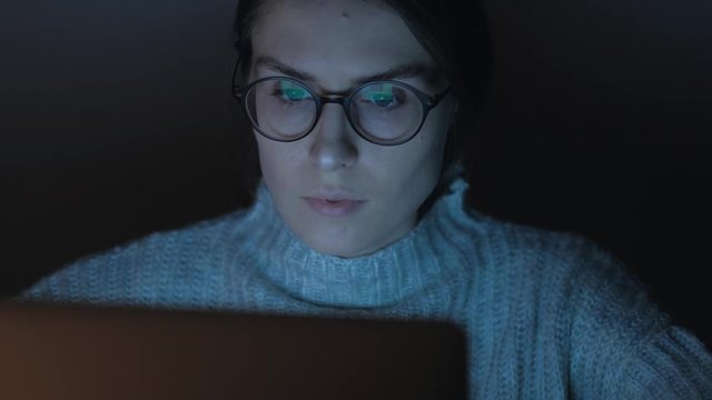 Panning shot - Portrait of a female working at the computer in the night, the reflection of the laptop screen with her glasses, woman browsing the Internet.