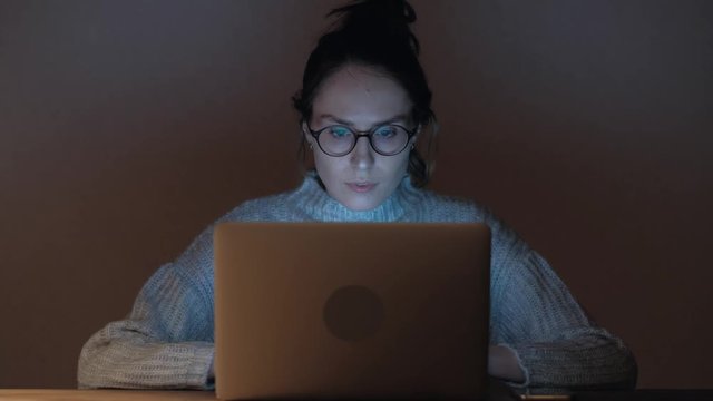 Cute woman in glasses browsing the Internet in dark room. 4k UHD. Portrait of a female working at the computer at night. 