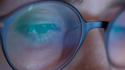 Extreme close up eye of woman working at laptop at night. 4k. Reflection of the laptop screen in her glasses.