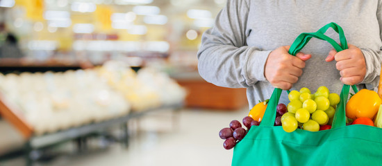 Customer hold reusable green shopping bag with fruit and vegetable over Supermarket aisle blur defocused product shelves interior bokeh light background
