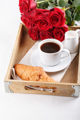 Cup of coffee and croissant on wooden tray. Festive breakfast with a bouquet of red roses on white table. Copy space.