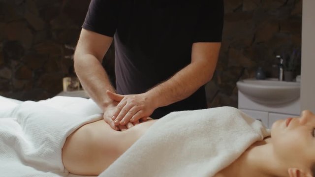 Male masseur burning fat by anti-cellulite massage. Young woman receiving stomach massage in spa, lying on massage table slide dolly camera movement