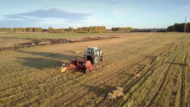 close tractor drives and rectangular baler pushes up collected straw bale on harvested field at sunset 
