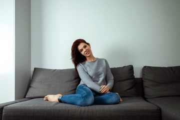 Fototapeta premium Young dreaming woman sitting on sofa at home. Thoughtful young woman sitting in living room and looking up. Smiling woman relaxing on couch in a modern home.