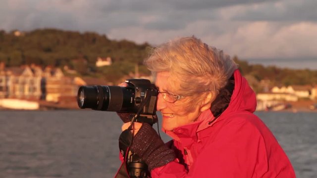 Happy senior woman in red jacket and gloves taking pictures with camera at sunset