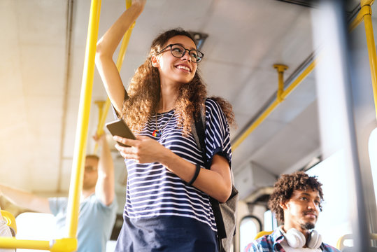 Beautiful mixed race girl with long curly hair using smart phone for reading or writing message while standing in city bus.