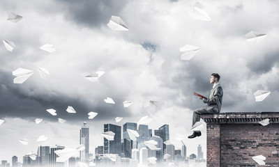 Man on roof edge reading book and cityscape at background