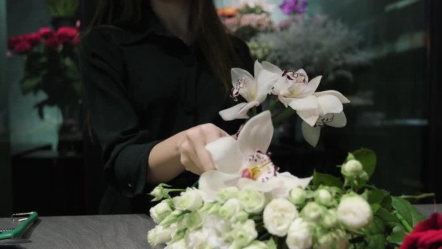 Professional florist works in a flower shop, prepare Orchid Cymbidium and white roses for bouquet, woman's hands close-up.