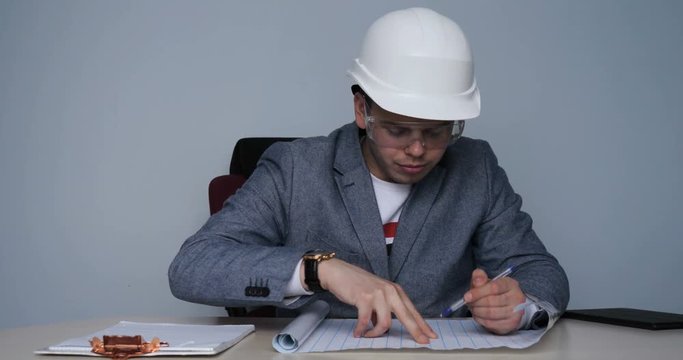 Young guy (male) An architect in a helmet and protective glasses makes up checking drawings in the office building. Concept of: Construction, Work, Lifestyle, In a jacket, Check.